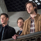 Sam and Dean look over a balcony while Castiel peeks over their shoulders.